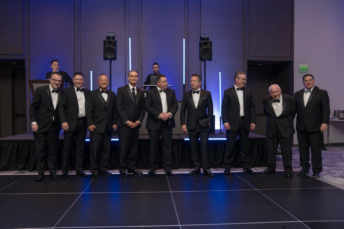 Group of men in formal tuxedos standing together on dark stage with blue lighting and speakers in background