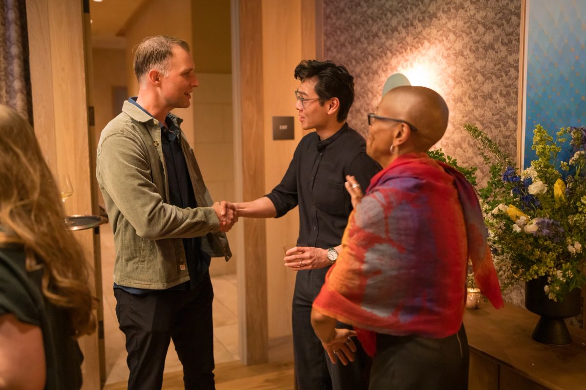 Three men shake hands and converse in an indoor setting with warm lighting and floral arrangements.
