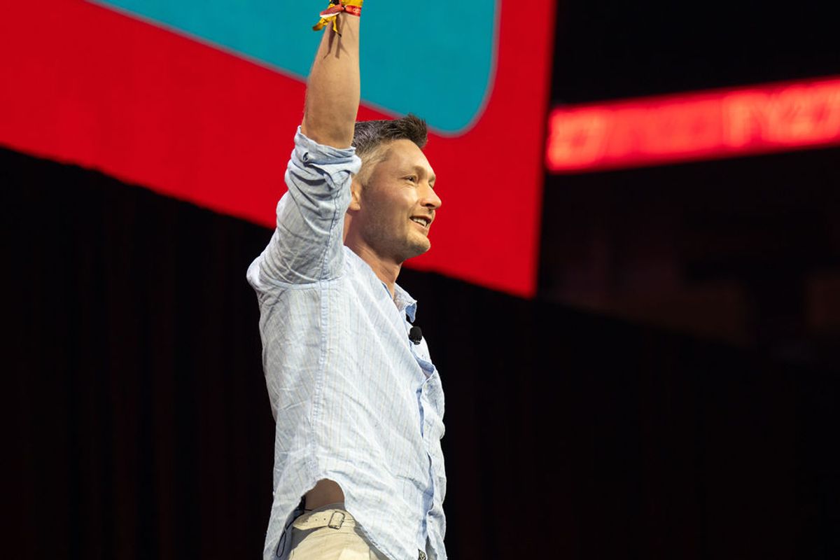 Speaker in light blue shirt raises arm while presenting on stage with red and teal backdrop at conference