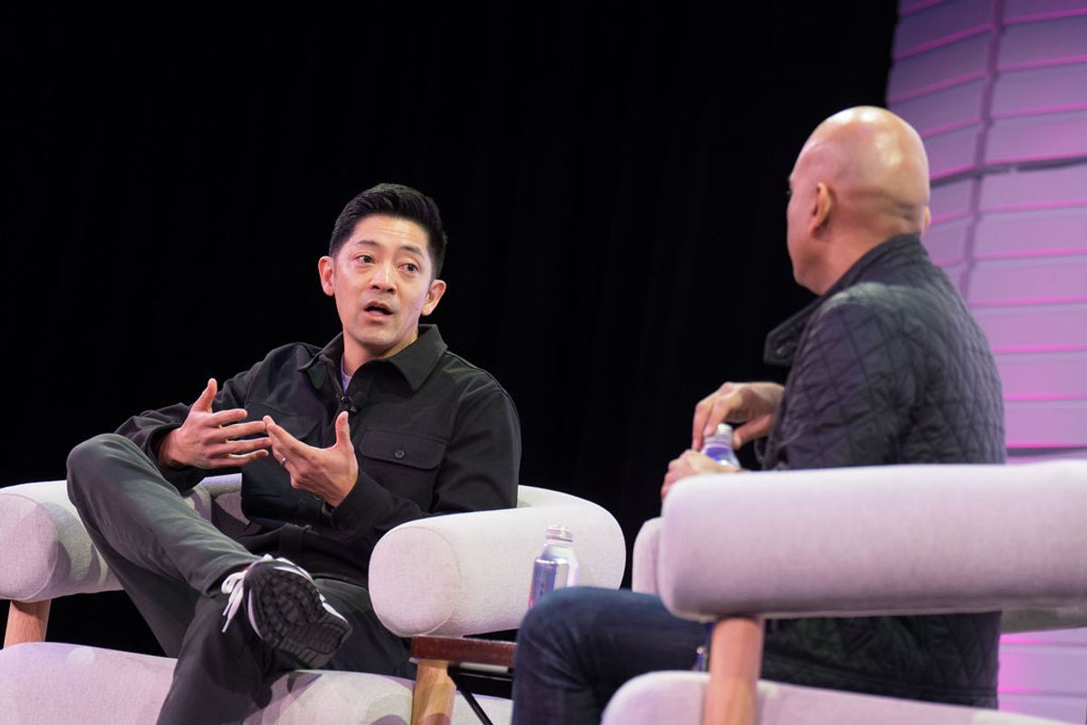 Two men in conversation on white chairs during a panel discussion, one gesturing while speaking to the other.