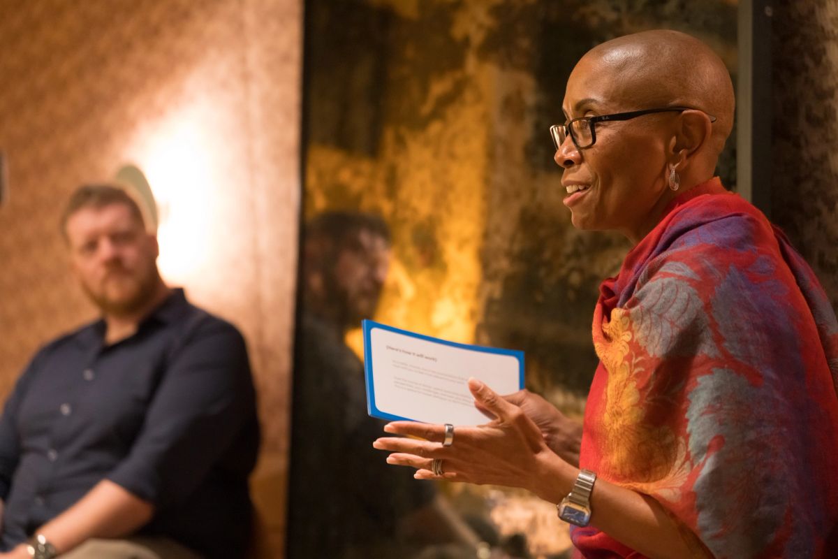Speaker with glasses and colorful shirt gestures while holding notes, with seated panelist in background