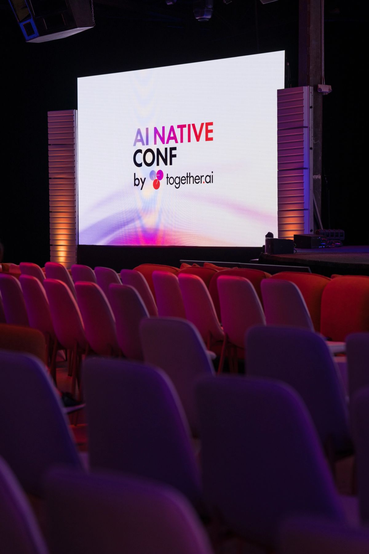 Conference auditorium with red seats facing large screen displaying "AI NATIVE CONF by together.ai" logo