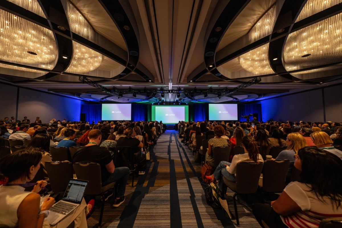 Large conference audience seated in ballroom facing three illuminated screens on stage.