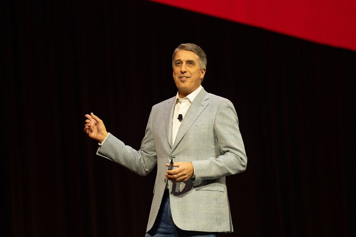Executive presenting on stage with microphone, wearing gray suit jacket, gesturing to audience against dark.