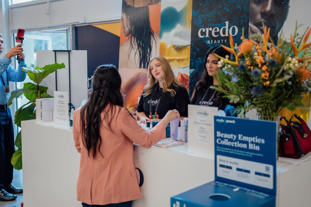 Two representatives at Credo Beauty booth interact with attendee at conference, with promotional materials.