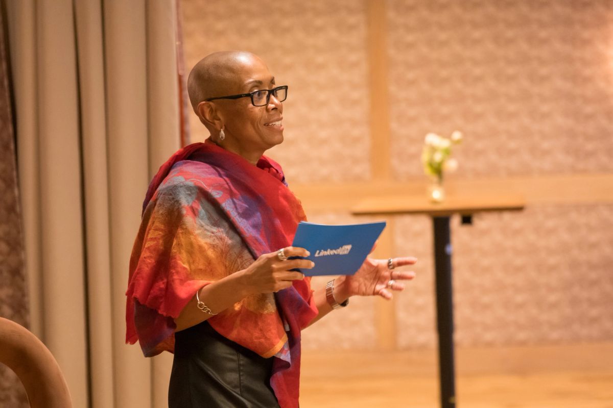 Speaker in colorful shawl holding blue folder addresses audience at panel discussion in brick-walled venue