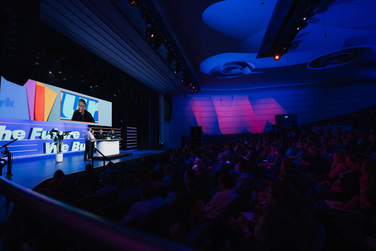 Speaker presenting on stage to seated audience in blue-lit auditorium with large screens displaying content