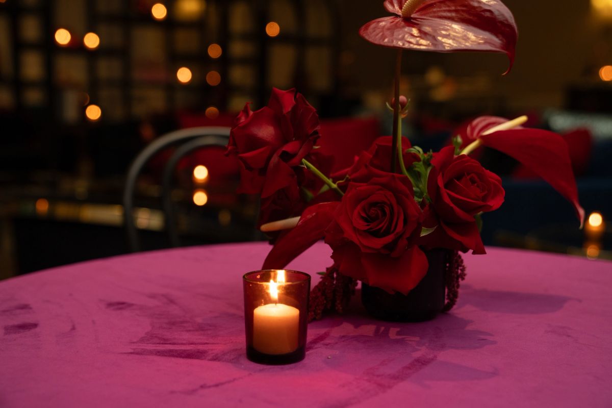Red roses and anthurium flowers in a dark vase with a lit candle on a purple tablecloth at an evening event.