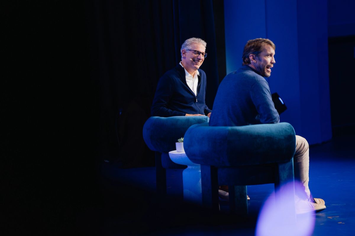 Two men sitting in blue armchairs on stage having a conversation at a conference event with blue lighting