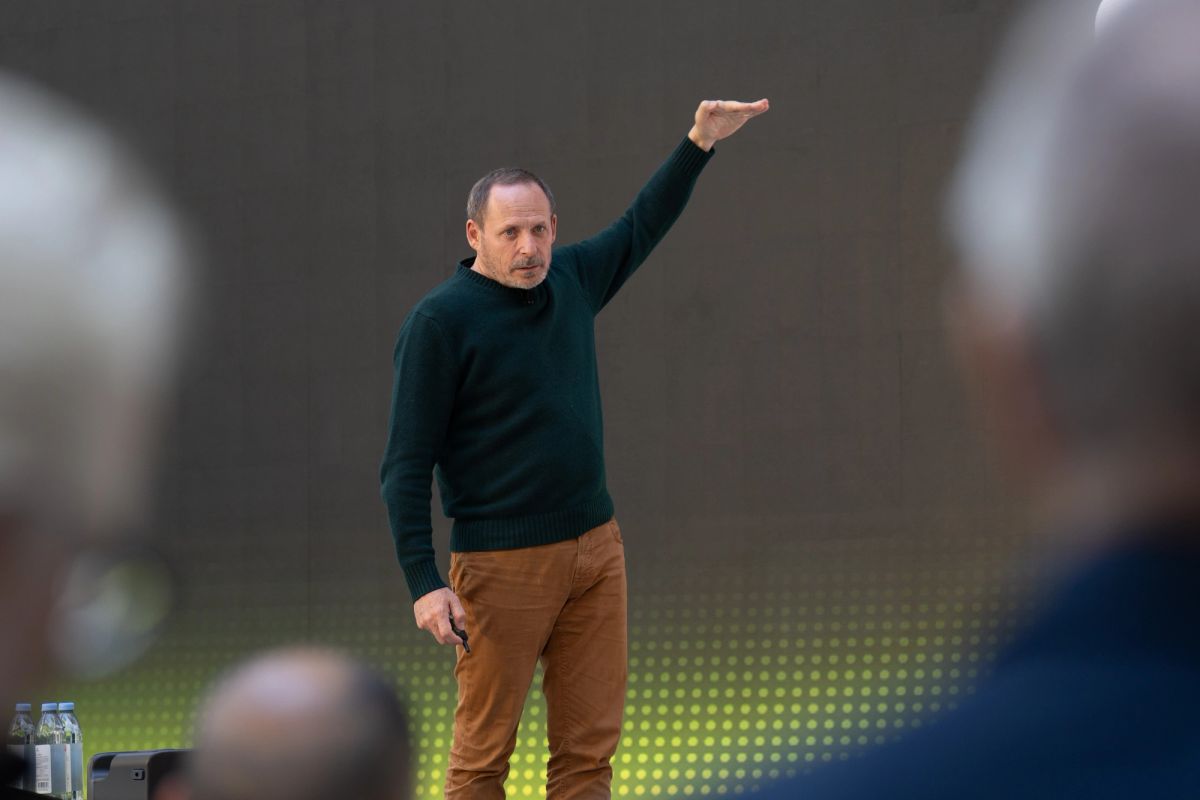 Speaker in green turtleneck raises arm while presenting to seated audience.