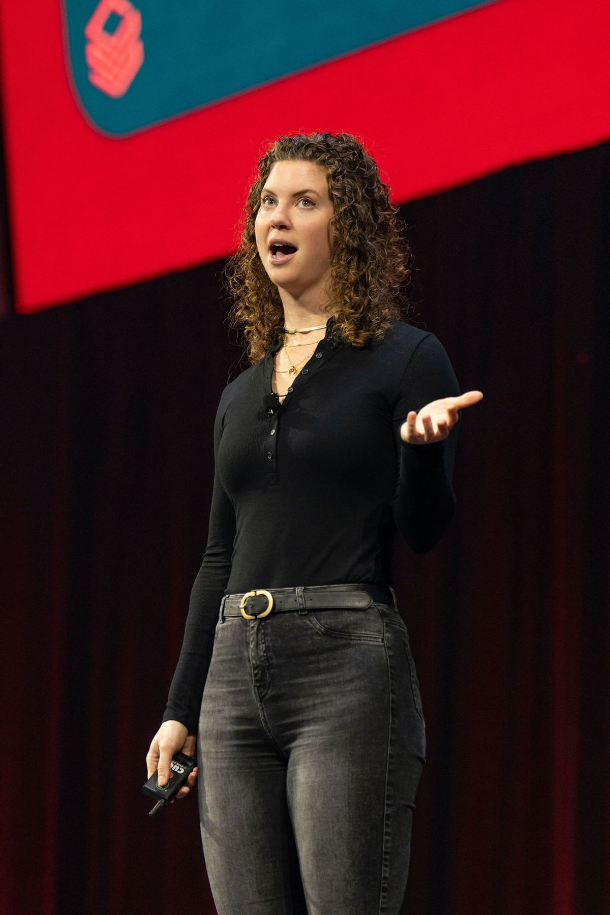 Speaker with curly hair presenting on stage with red backdrop, gesturing with one hand while holding device
