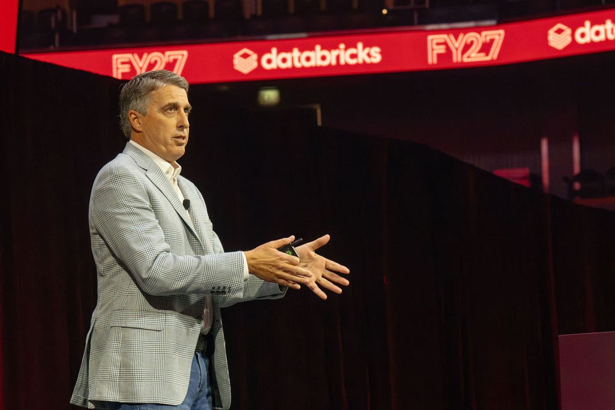 Speaker in gray blazer gesturing while presenting on stage with red Databricks and FY27 signage visible