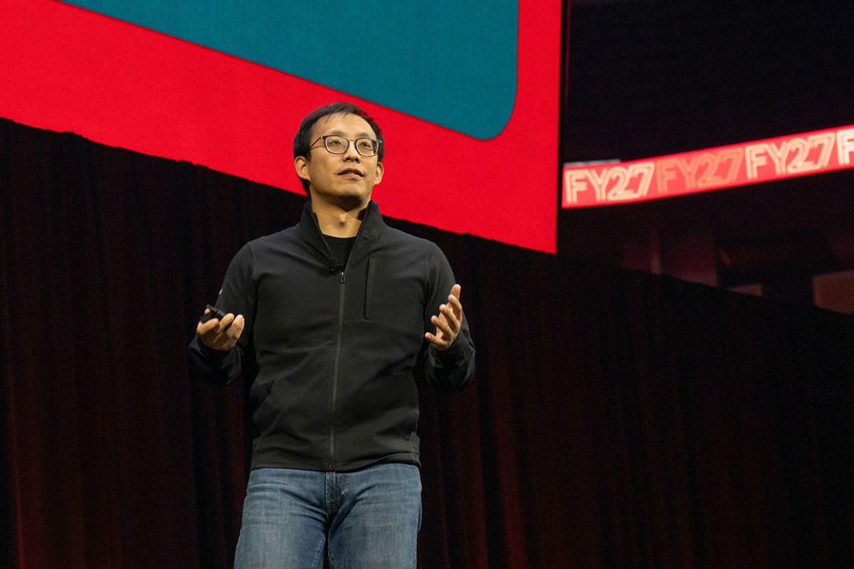 Speaker presenting on stage with red and teal geometric backdrop at conference venue