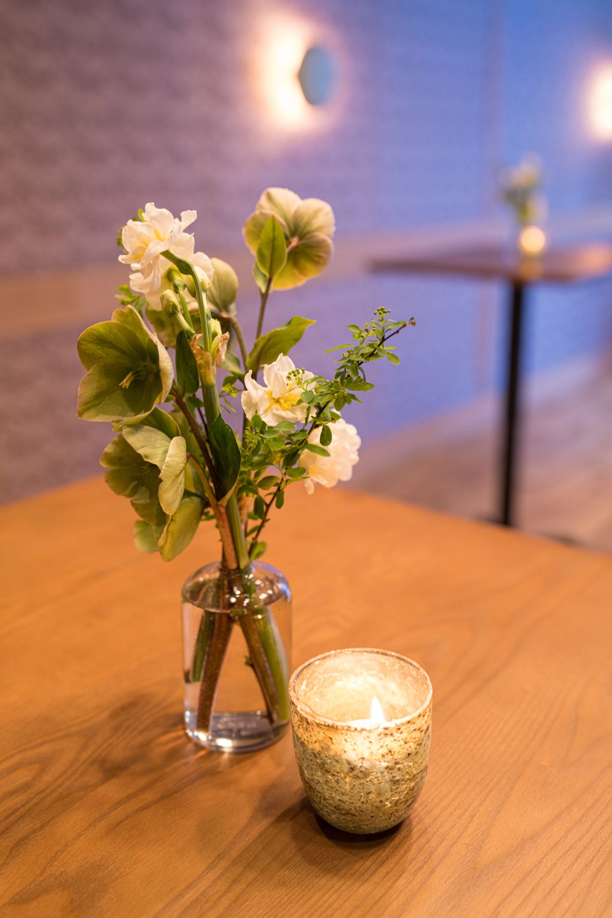 Small glass vase with white flowers and greenery sits on wooden table next to lit candle in textured holder.