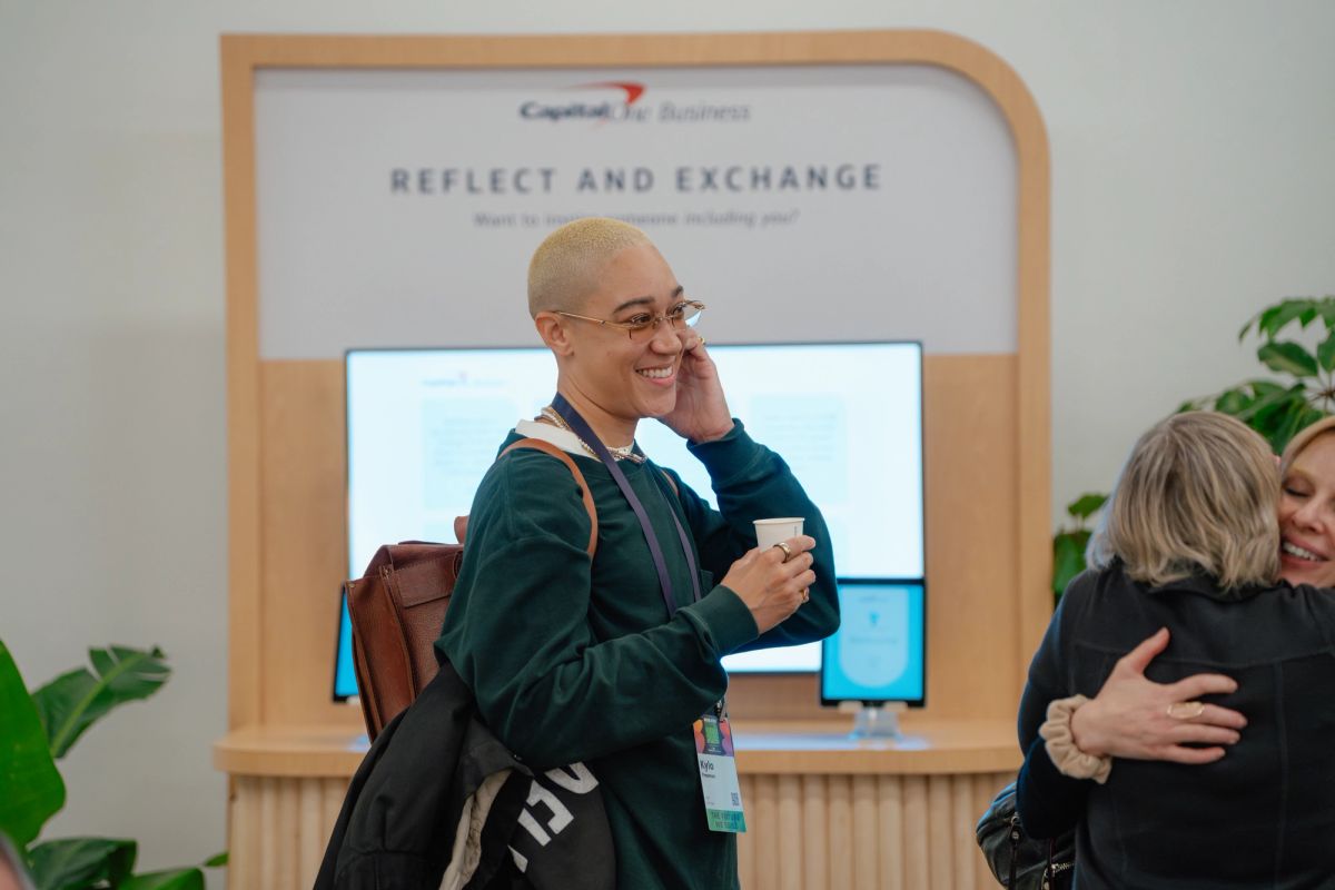 Conference attendee on phone while two people hug at networking event with "Reflect and Exchange" backdrop
