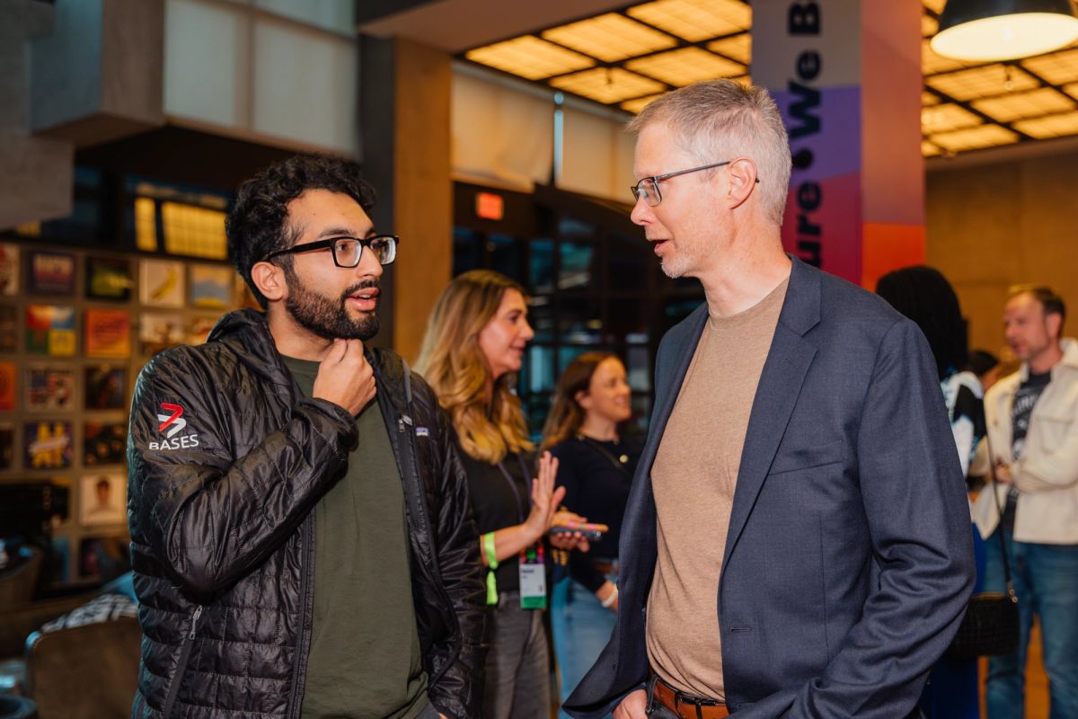 Two men in conversation at a conference networking event, with other attendees mingling in the background