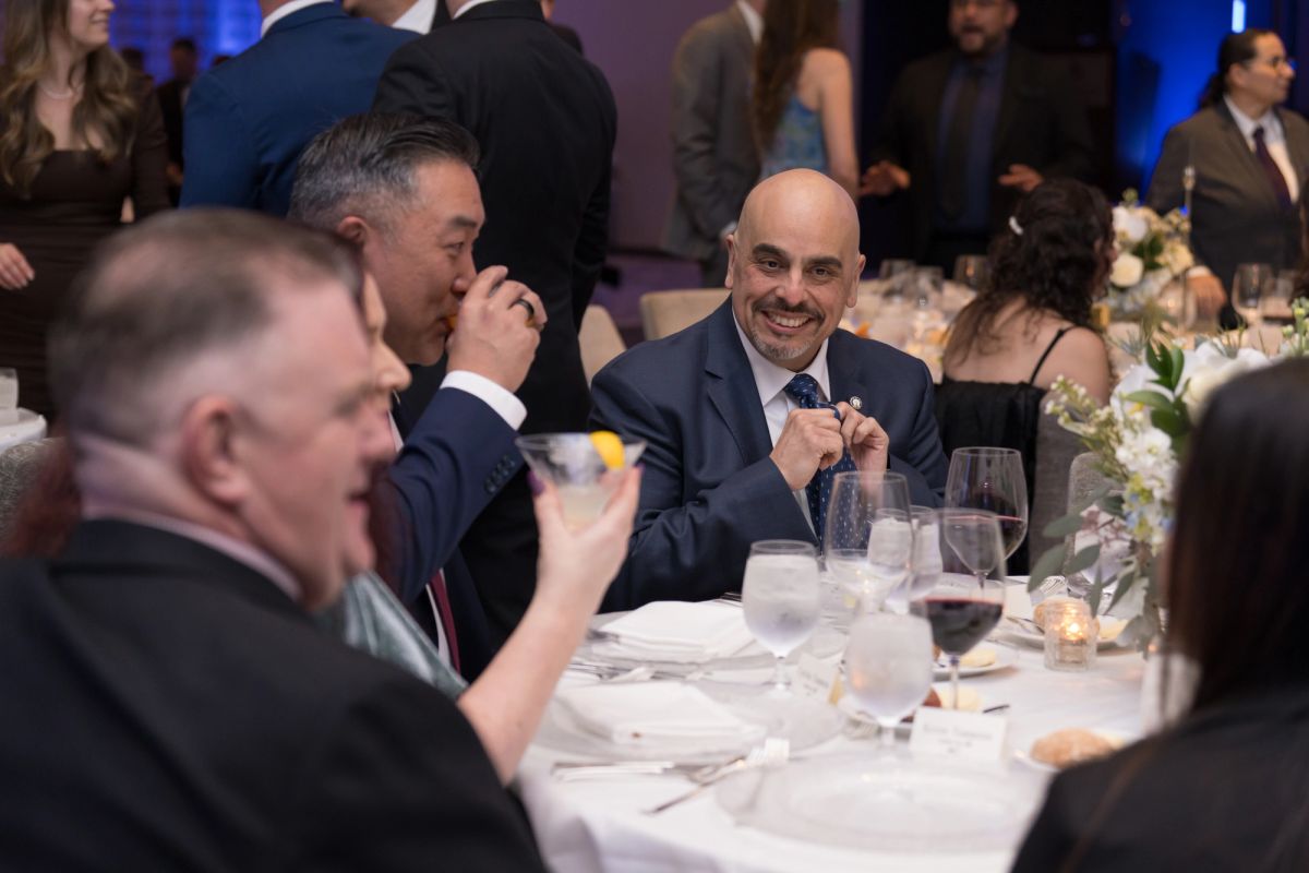 Business executives in suits laughing and conversing at formal dinner table with wine glasses and floral.