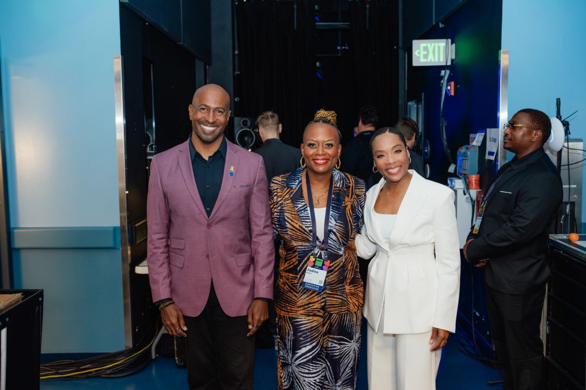 Three people pose together backstage at an event venue, with an exit sign visible in the background.