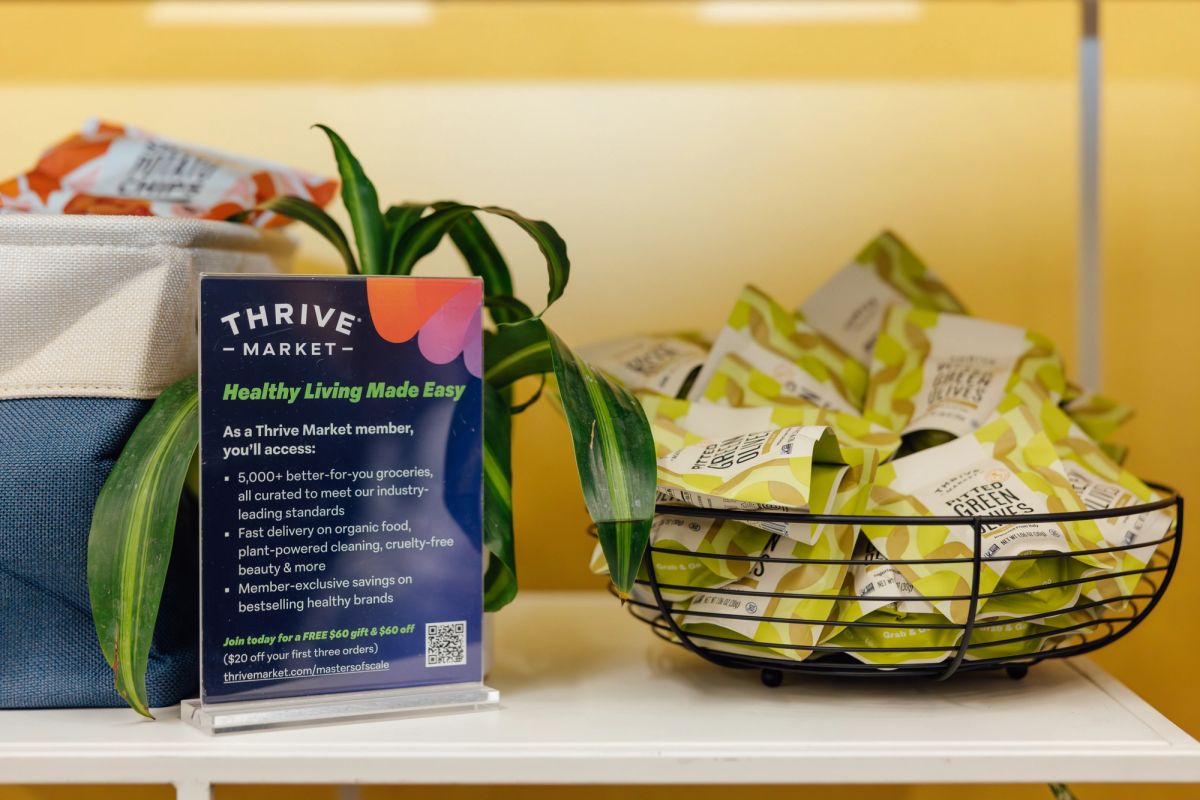 Thrive Market promotional card next to wire basket of green packages and plant on white shelf