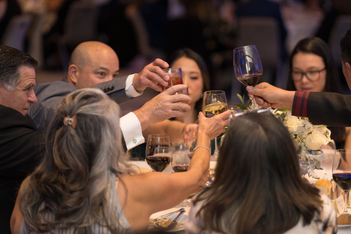 Guests raise wine glasses in a toast at a formal dinner event with floral centerpieces and attendees seated.