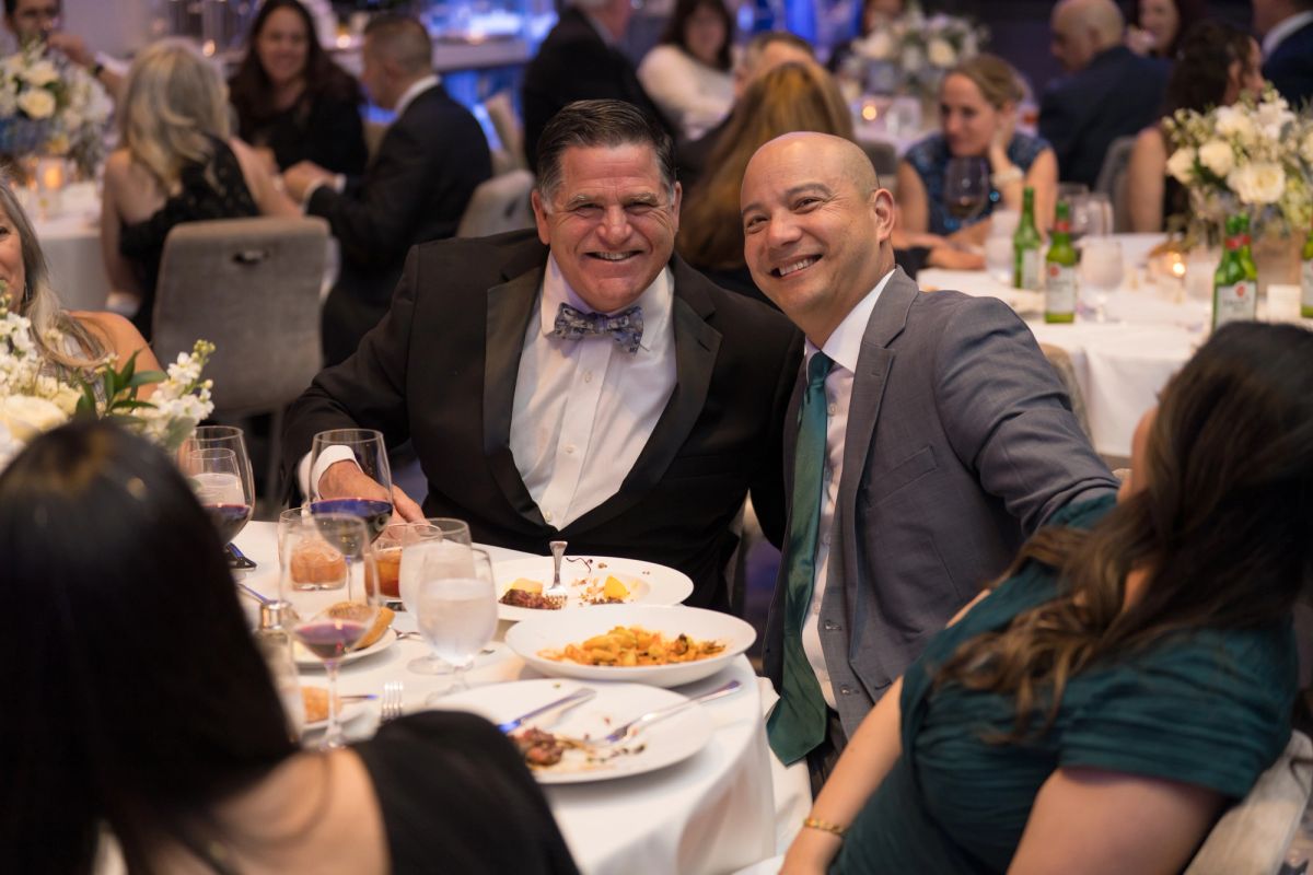 Two men in formal attire smile at camera while seated at dinner table during formal gala event