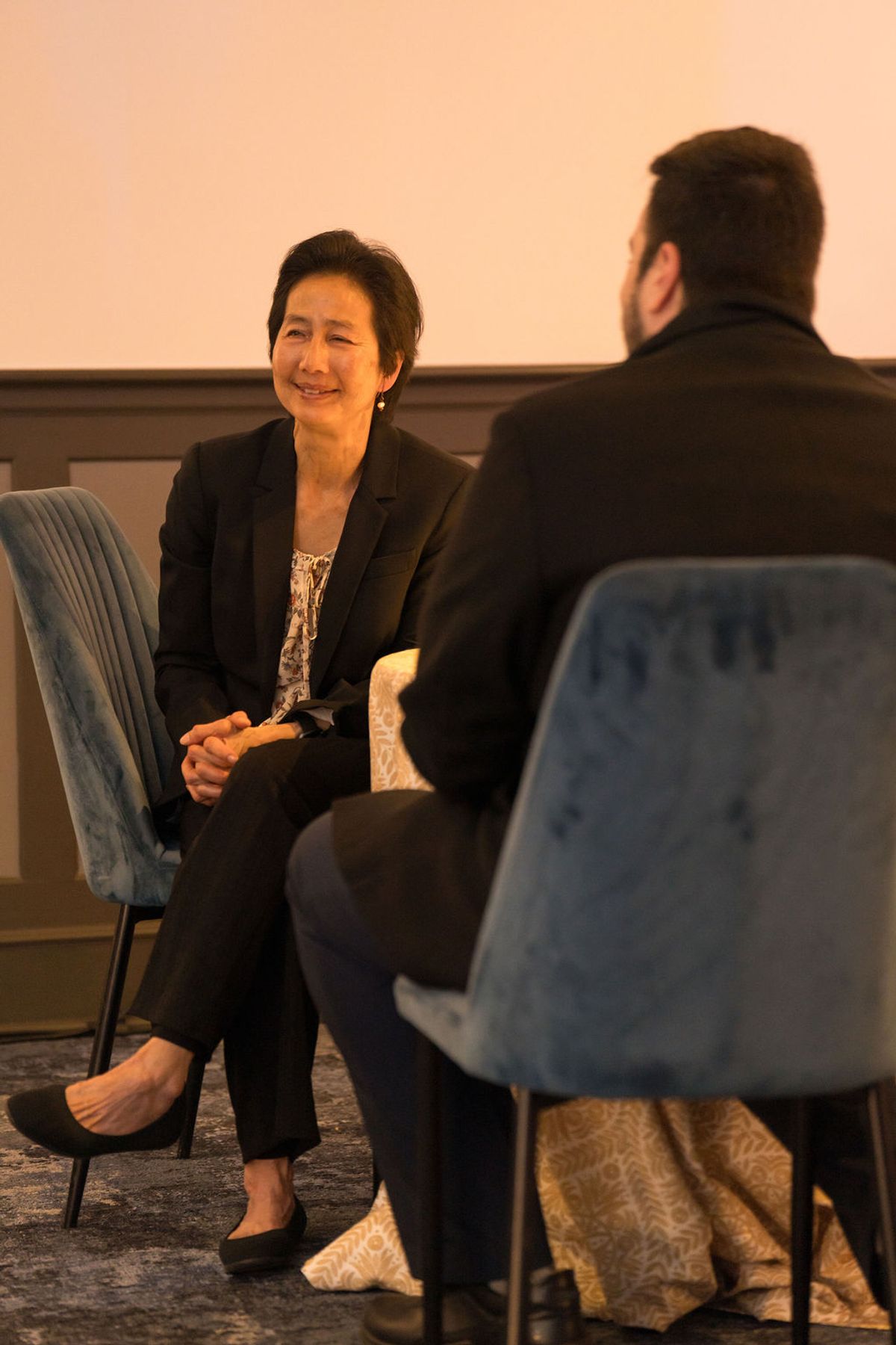 Two business professionals in conversation, woman in dark suit seated in blue chair facing man