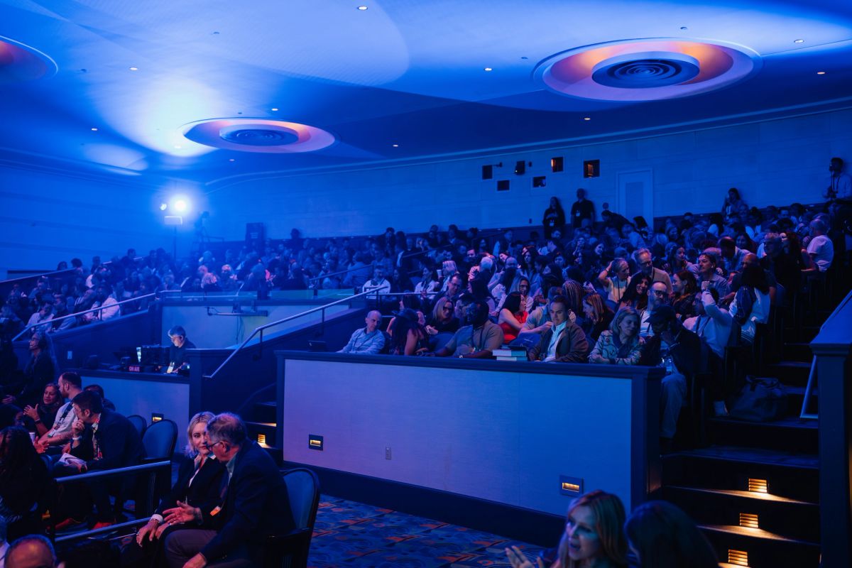 Audience members seated in tiered seating at a conference venue with blue ambient lighting and circular.