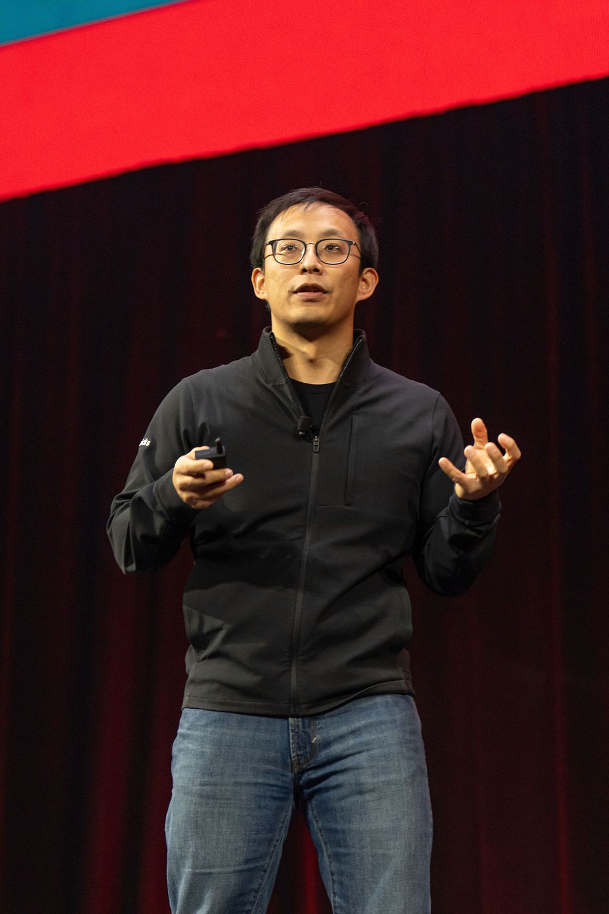 Speaker presenting on stage holding smartphone, wearing glasses and dark zip-up jacket, with red and teal.