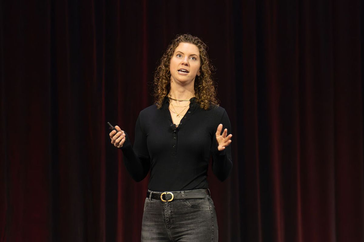 Speaker with curly hair presenting on stage, holding phone and gesturing with one hand against dark curtain.