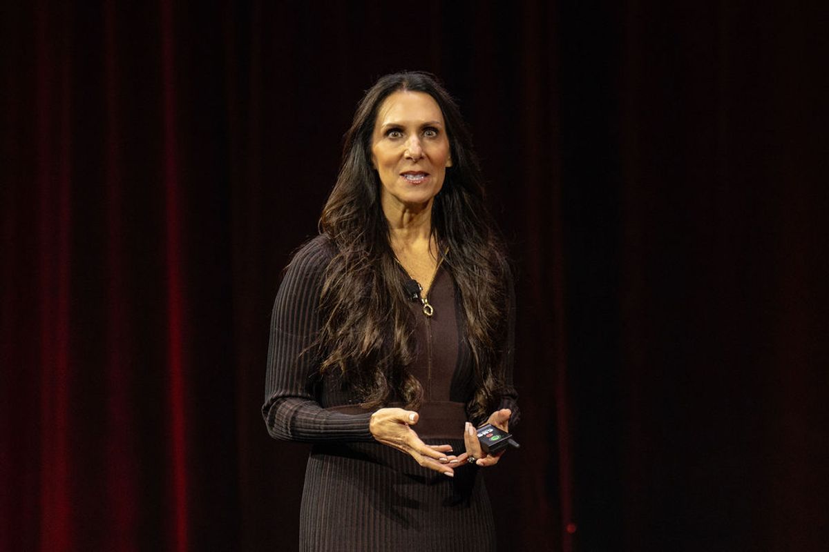 Speaker with long dark hair presenting on stage, wearing black ribbed sweater, holding mobile device