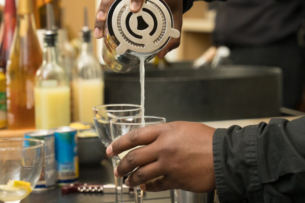 Bartender straining a cocktail into a glass at a bar with bottles and supplies in the background