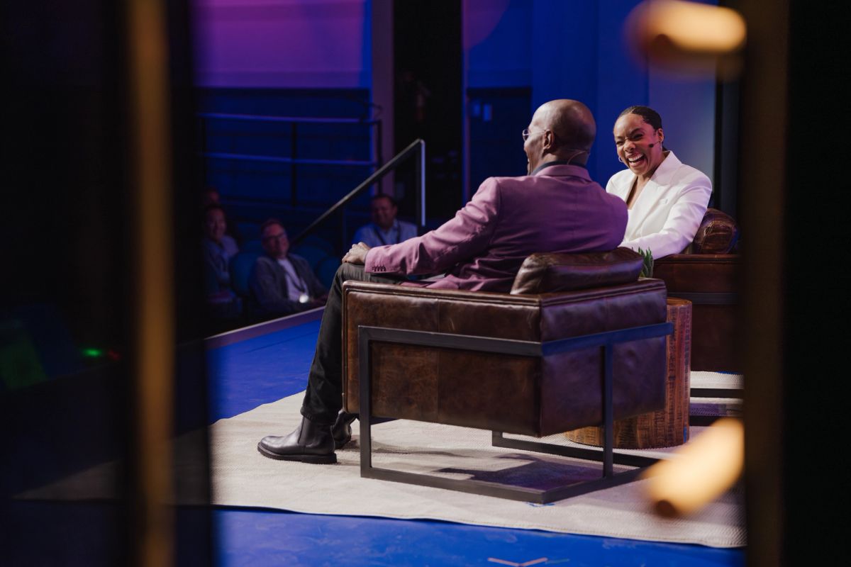 Two people sitting on a couch having a conversation on a lit stage with audience members visible in the.