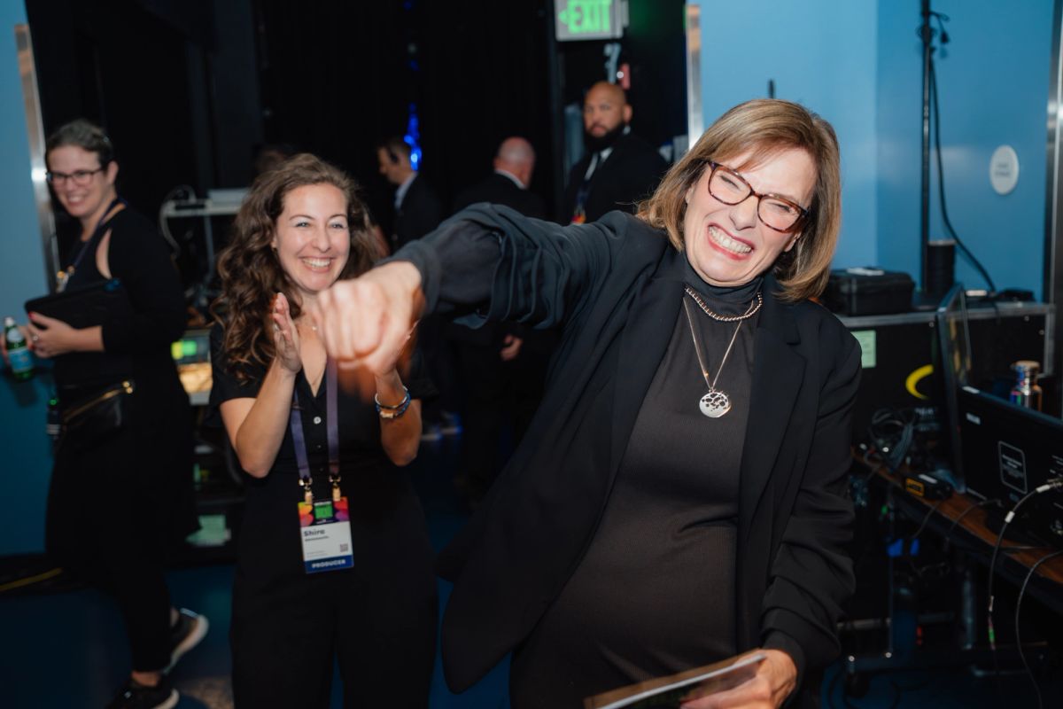 Two women celebrating and fist-bumping in a theater setting with stage equipment and attendees visible in.
