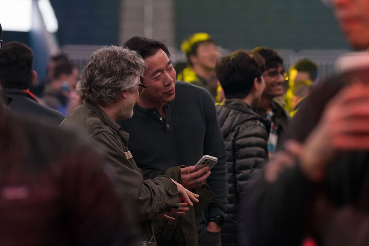 Two men in conversation at a conference networking event, with other attendees visible in background