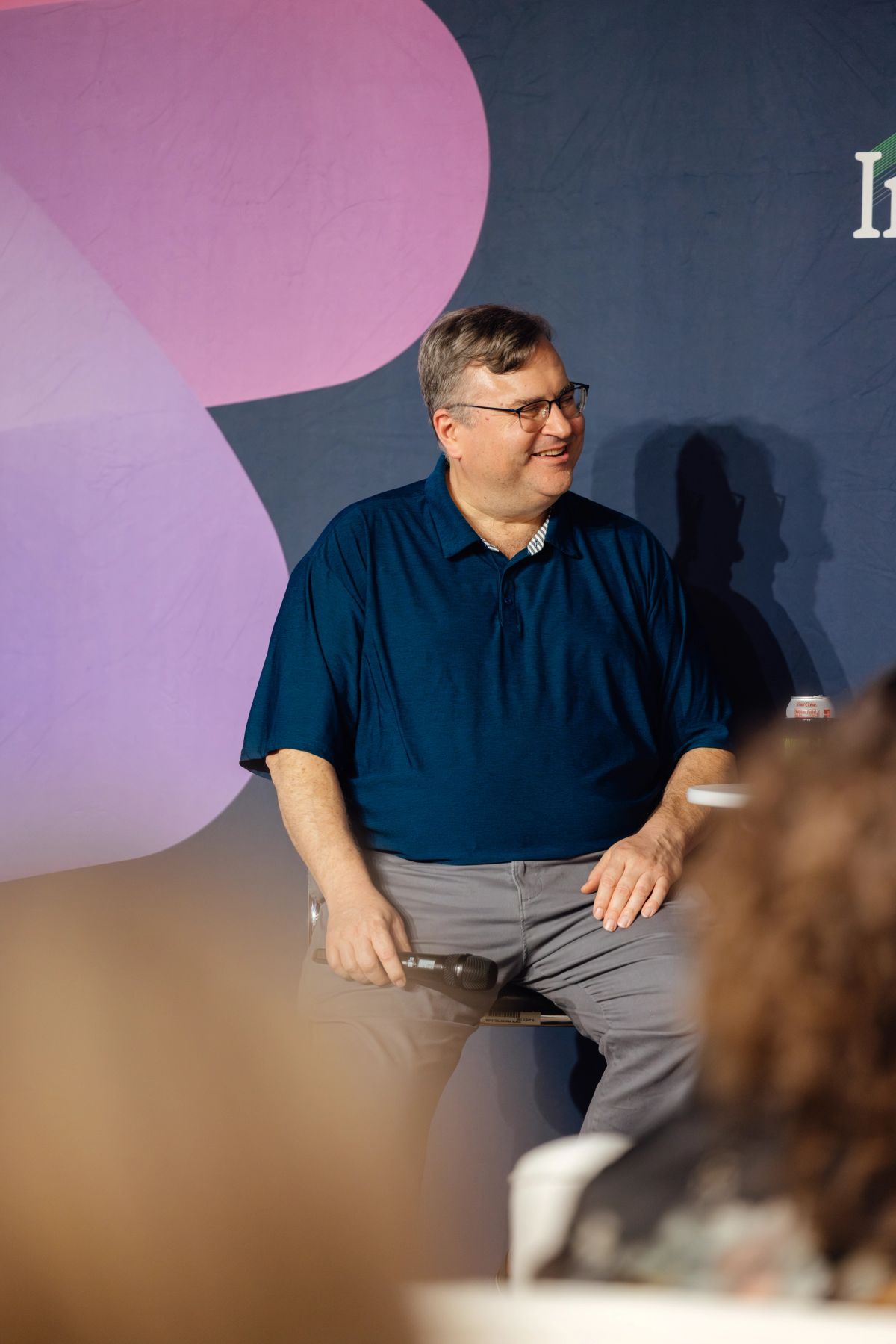 Speaker in blue polo shirt sits smiling on stage with purple and blue backdrop at conference event