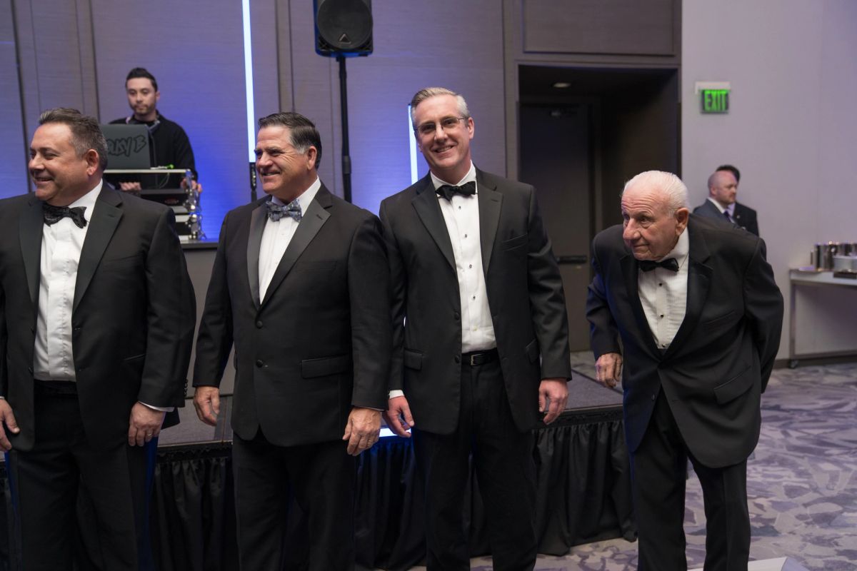 Four men in black tuxedos and bow ties standing together at a formal gala event in an elegant venue.