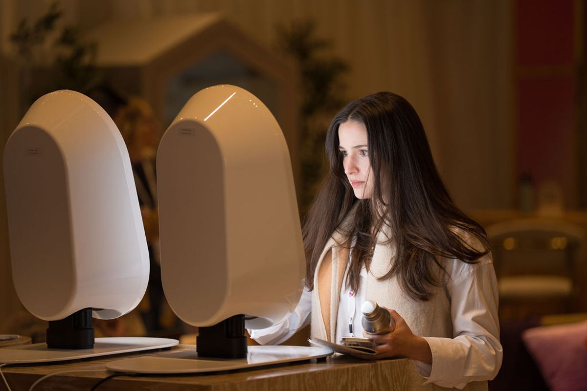 Woman with long brown hair sits at wooden table with two modern white lamps in warmly lit interior space.