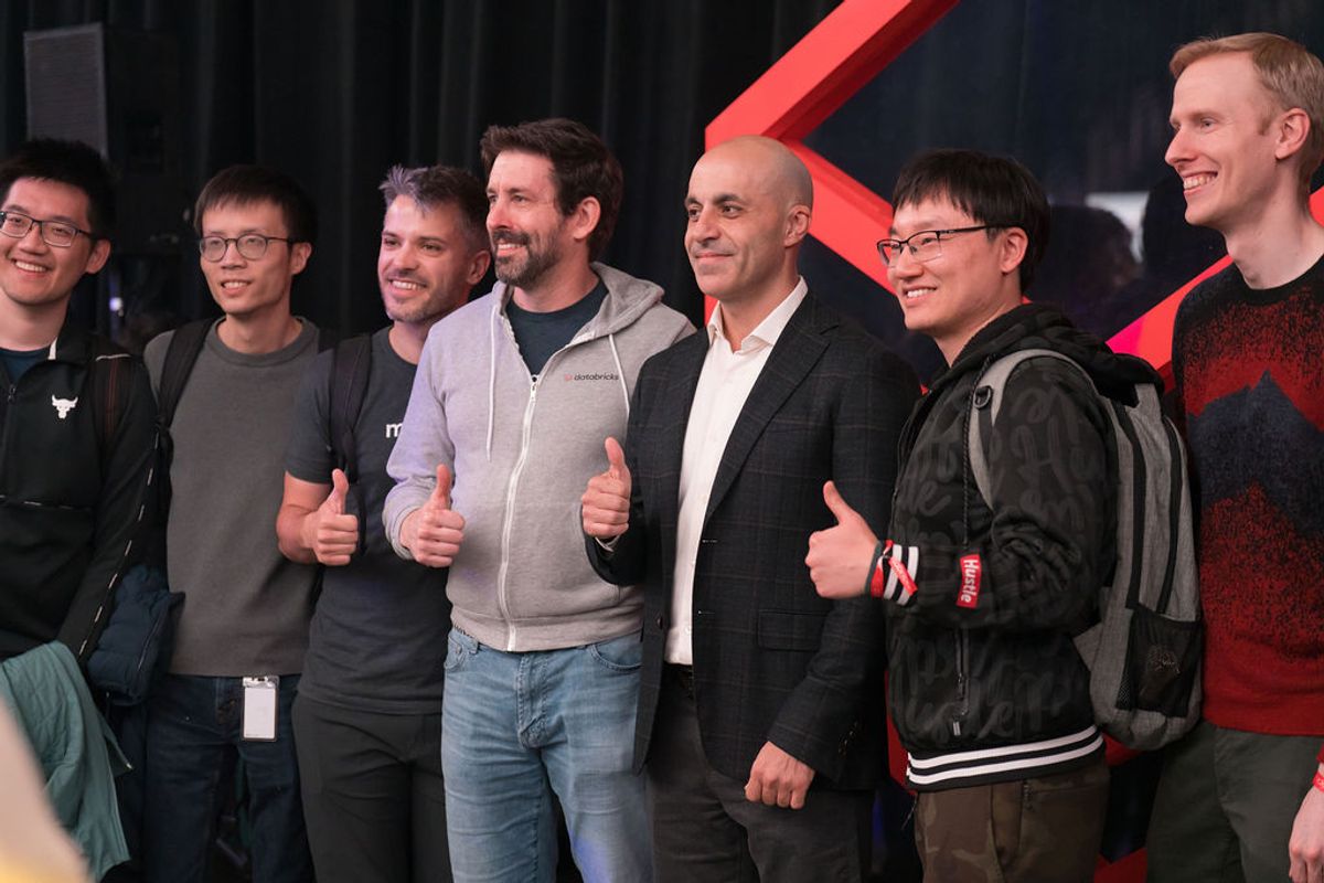 Seven men standing together on stage giving thumbs up gestures, smiling at audience during conference event
