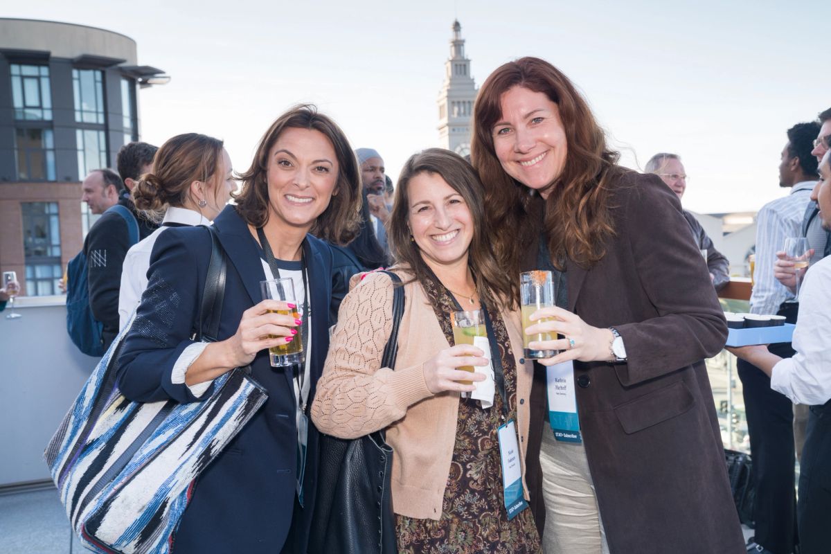Three women networking with drinks at the rooftop reception with the San Francisco skyline behind them