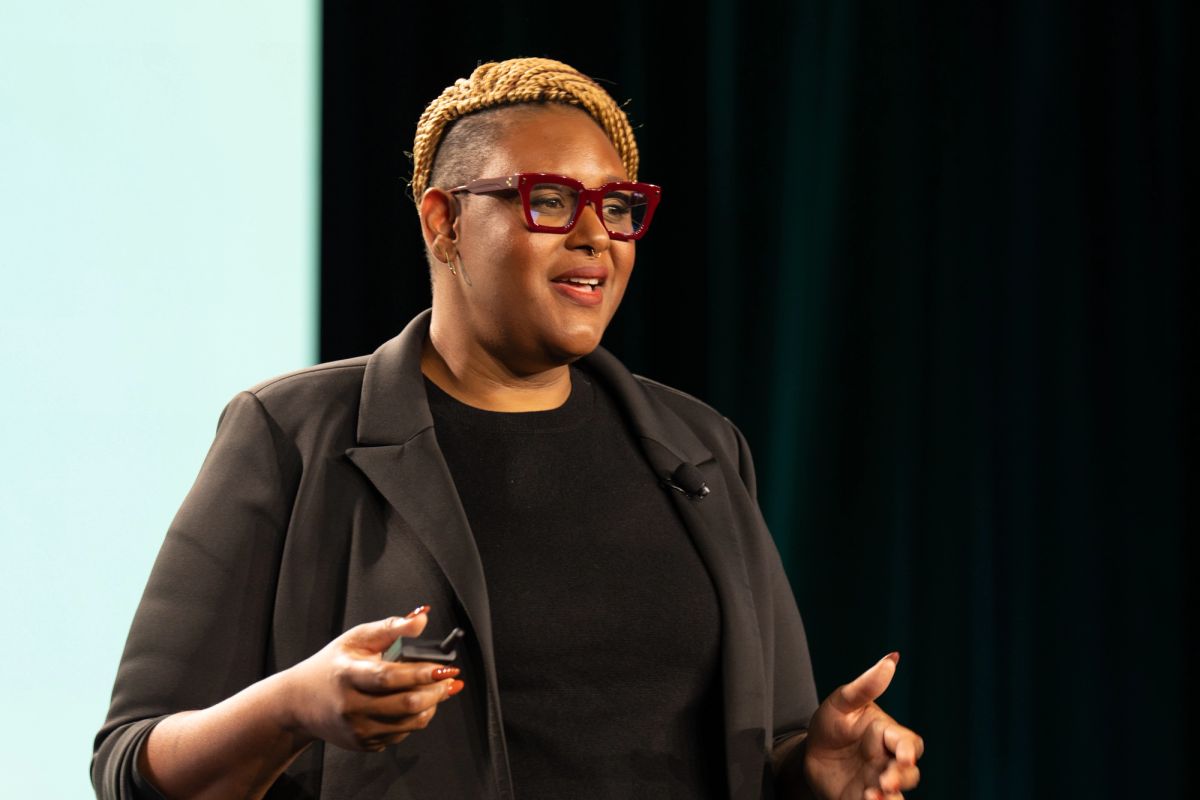 Speaker presenting on stage, gesturing while holding a clicker, wearing red glasses and black blazer.