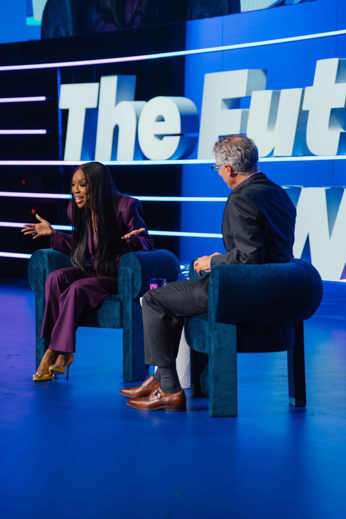 Two speakers in conversation on stage, woman in purple suit gesturing, man in dark suit listening, blue.