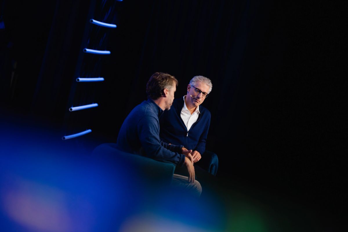 Two men in business attire having a conversation on stage with blue lighting and architectural elements in.