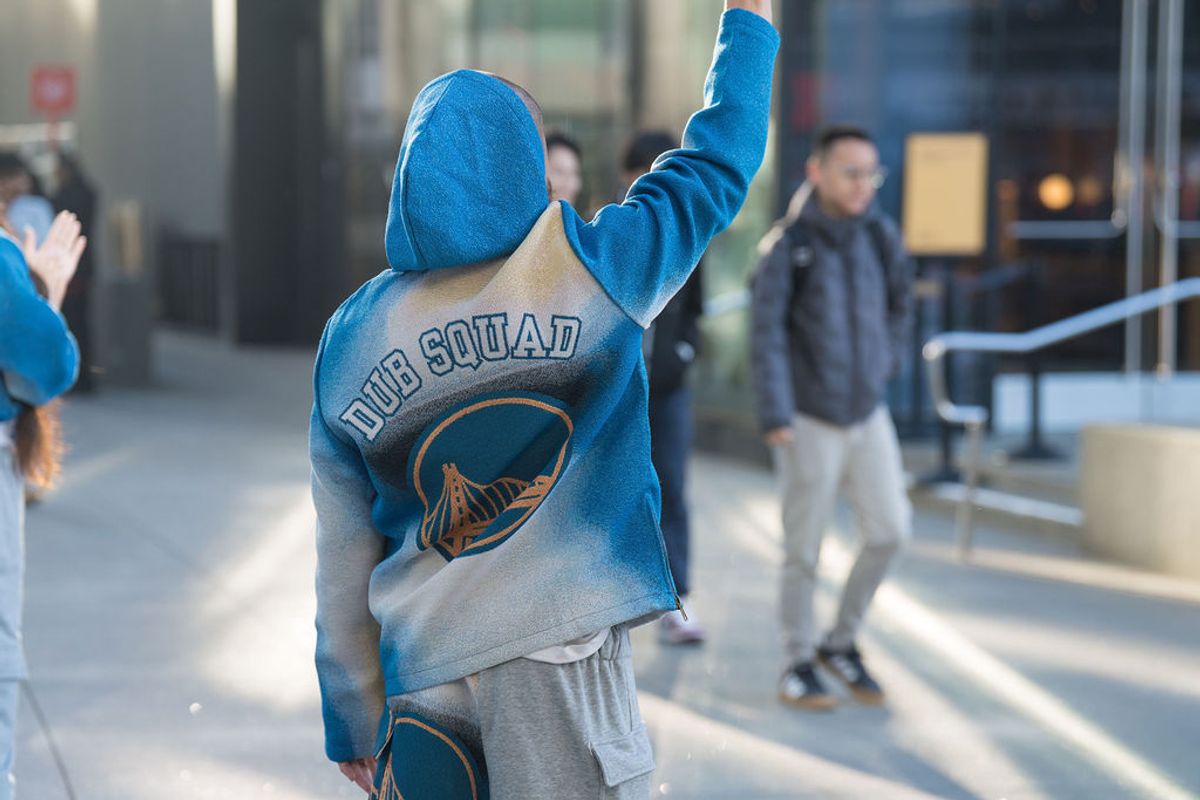 Person in blue Dub Squad hoodie with raised arm walking on outdoor steps near building entrance