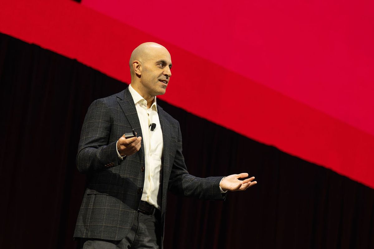 Bald speaker in gray suit presenting on stage with red and black backdrop at conference