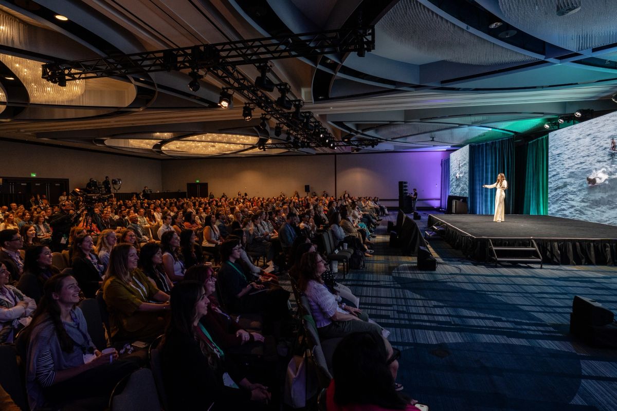 Speaker on stage addresses large seated audience in a conference ballroom with projection screen.