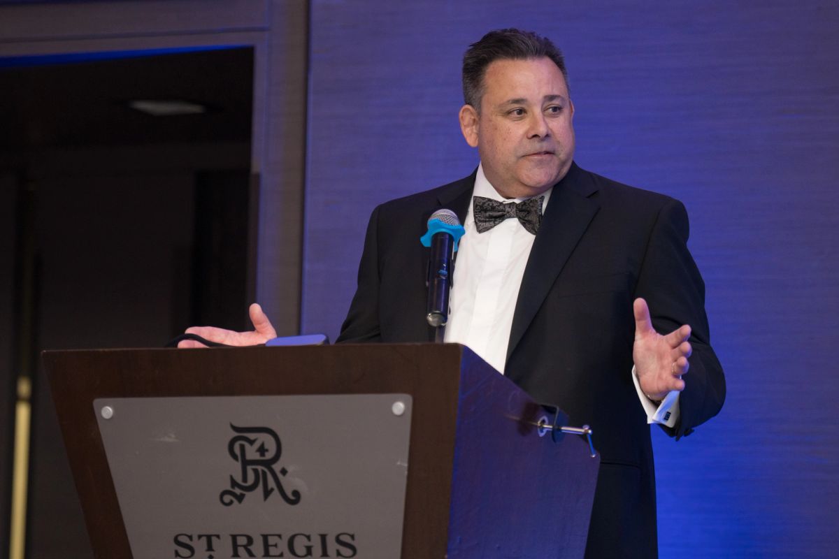 Formally dressed speaker in tuxedo gesturing at podium with microphone at St Regis hotel event