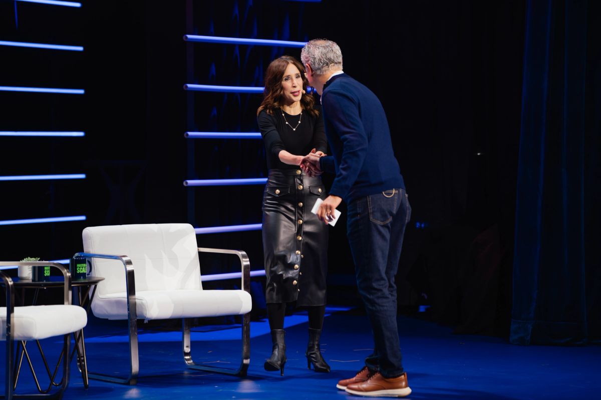 Two speakers having a conversation on stage with white chairs and blue lighting at a conference venue.