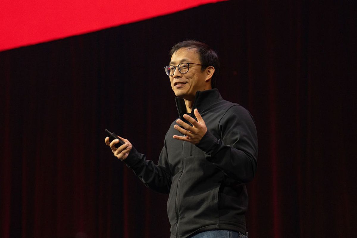 Speaker in dark jacket gesturing while holding phone on stage with red and black backdrop