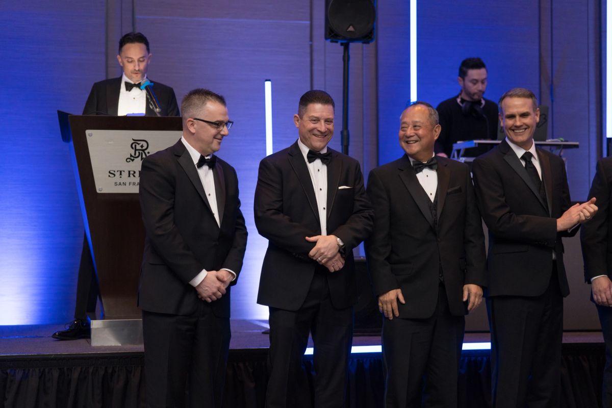 Speaker at podium addresses four men in tuxedos on stage at formal gala event with blue lighting