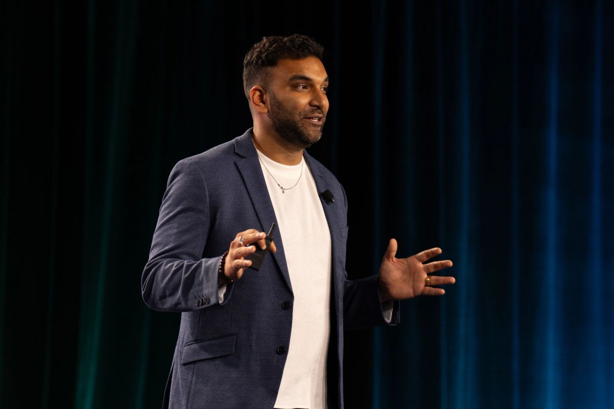 Speaker gestures while presenting on stage before a teal curtain backdrop.
